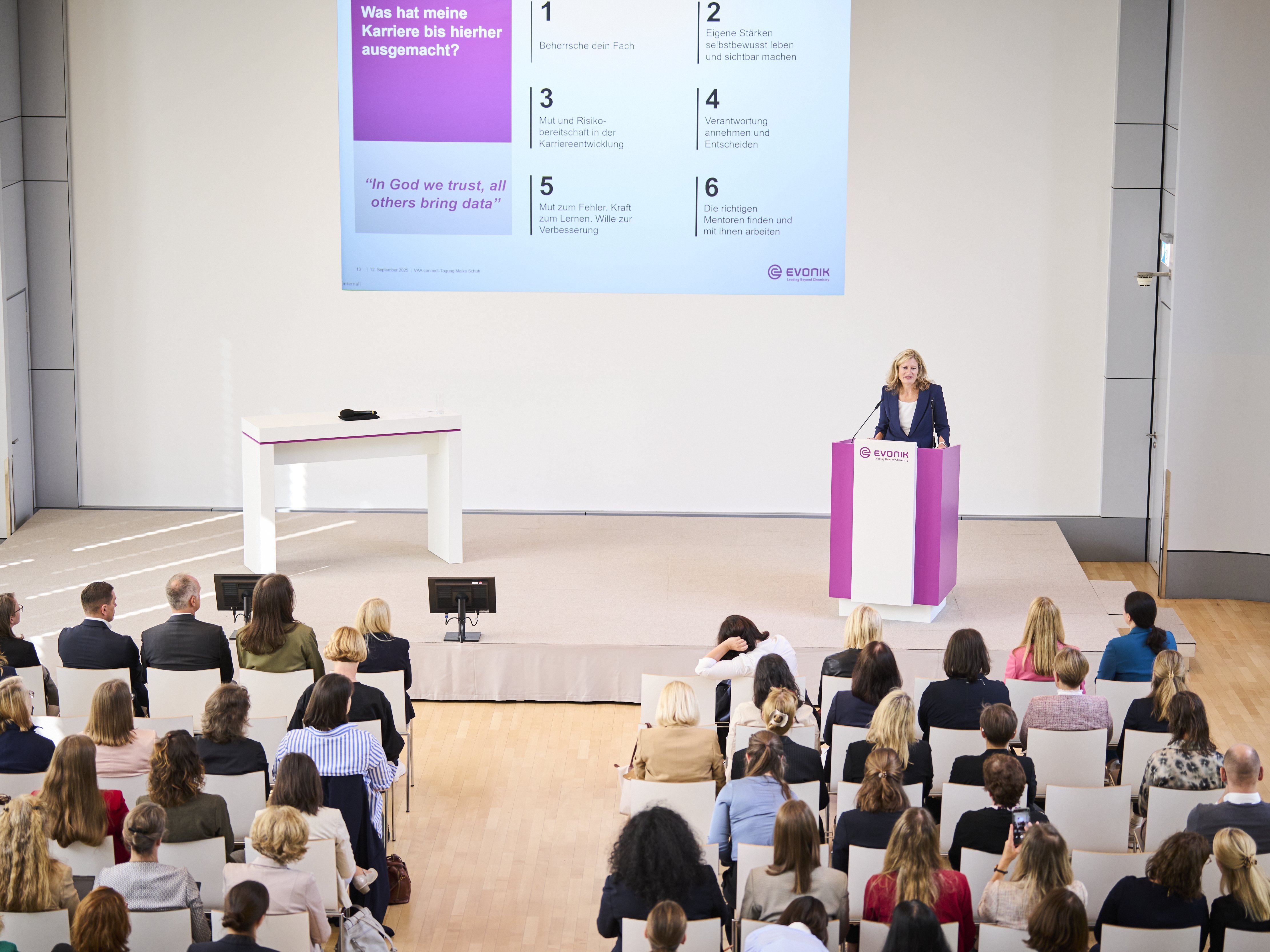 Der Saal von oben: Teilnehmerinnen und Teilnehmer schauen auf die Bühne, wo Maike Schuh steht und spricht.