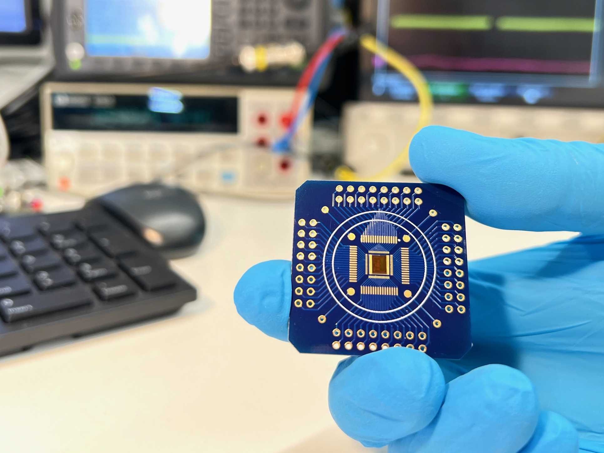 Eine behandschuhte Hand hält einen Chip. Im Hintergrund ein Schreibtisch mit Tastatur, Computer und Messgeräten.