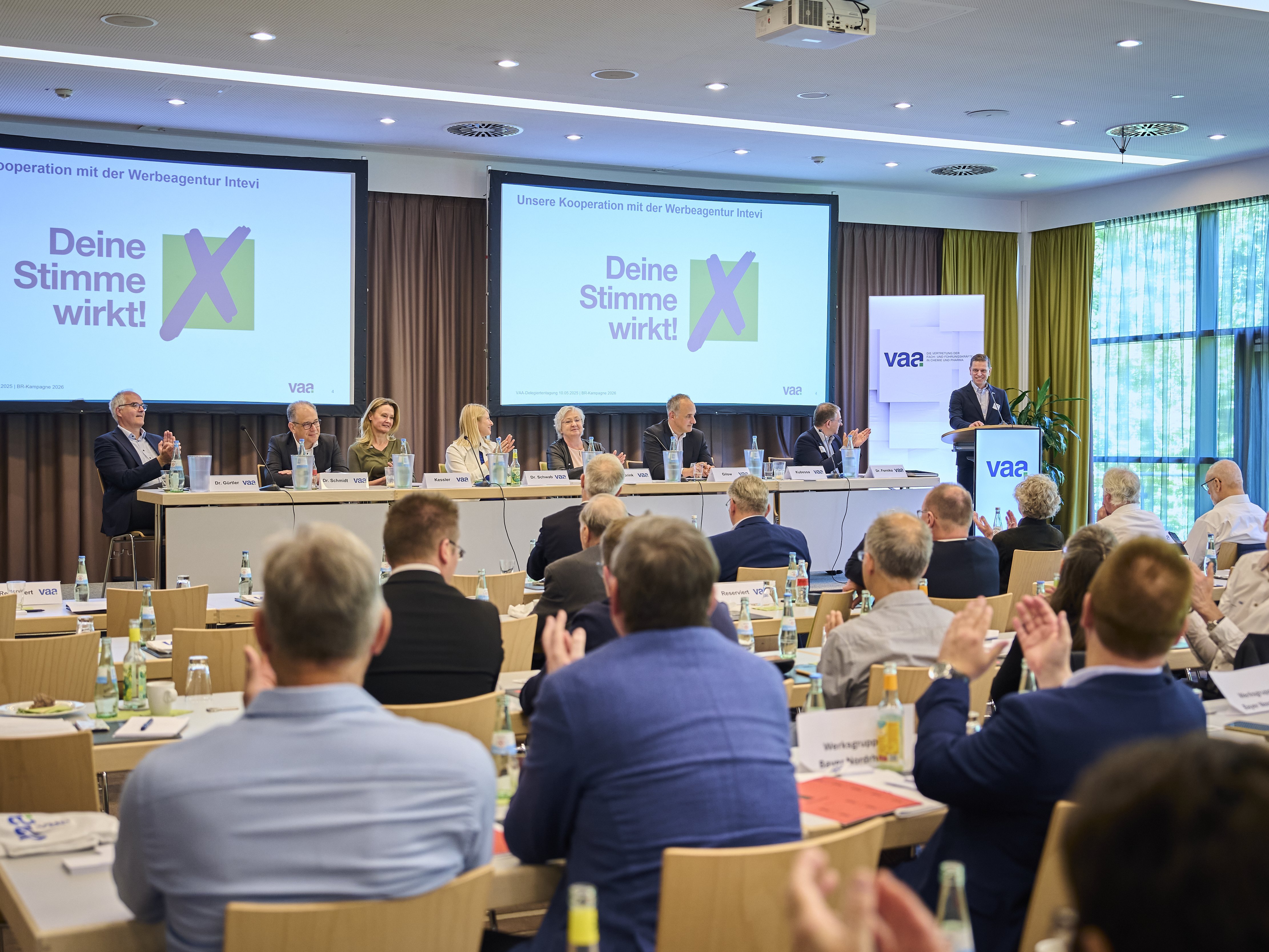 Applaudierendes Plenum von hinten, während Martin Kubessa das Betriebsratswahllogo vorstellt. Zu sehen auf den Leinwänden.