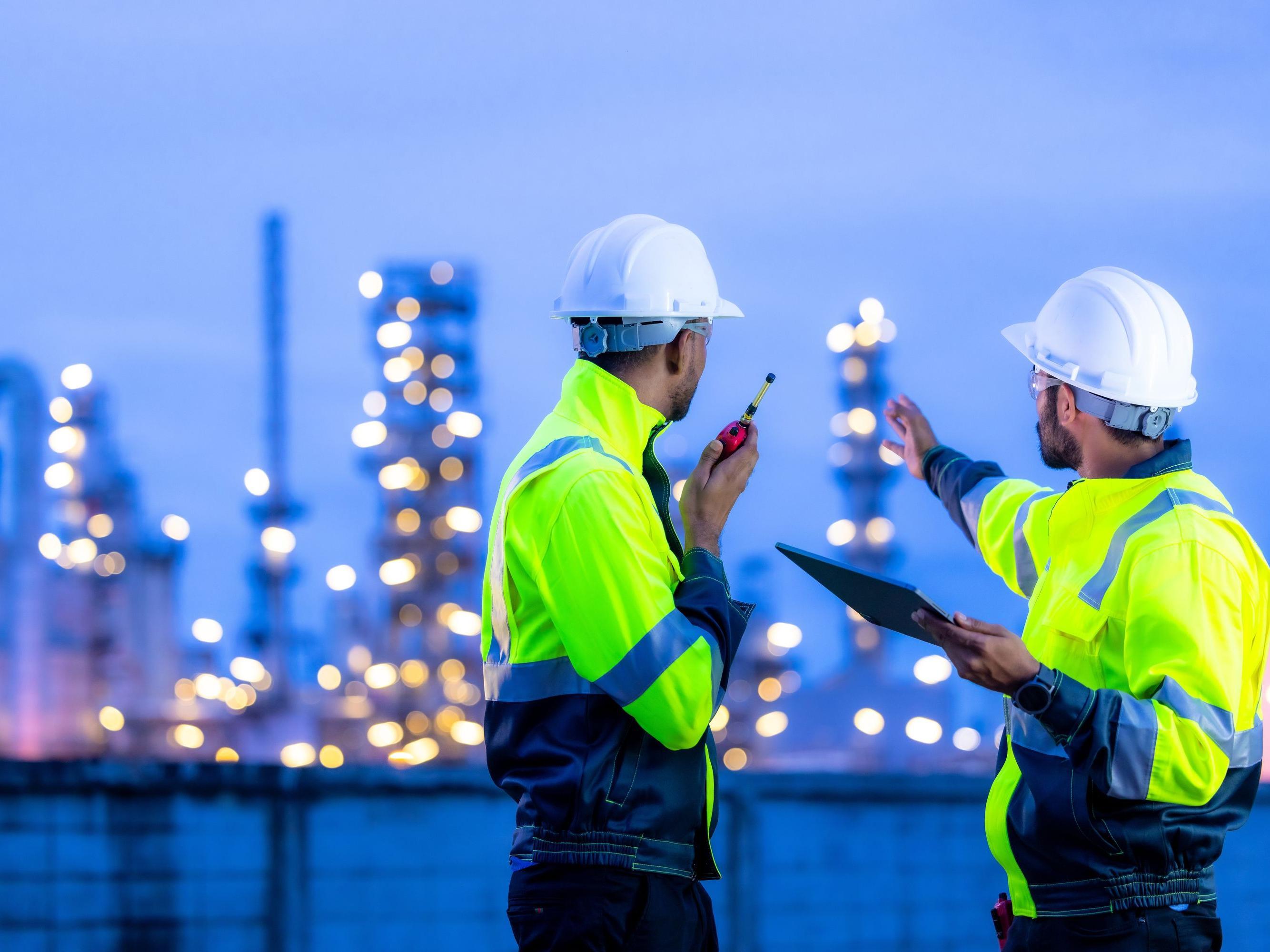 Zwei Industriebeschäftigte in gelben Overalls und mit weißen Helmen schauen auf ein großes Chemiewerk im Hintergrund.