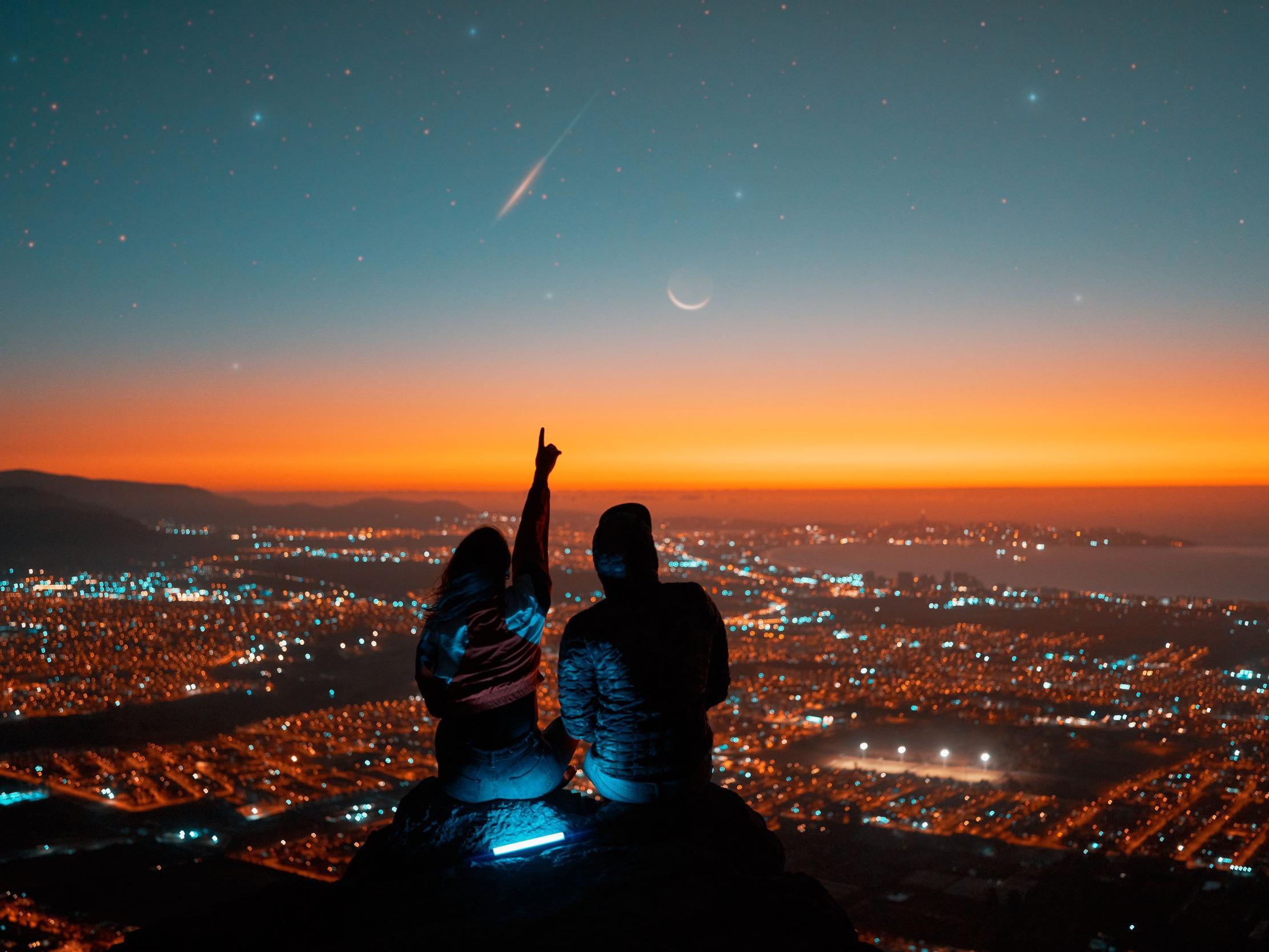 Ein Pärchen sitzt spätabends auf einem Berghang über einer Großstadt und schaut in den Sternenhimmel.