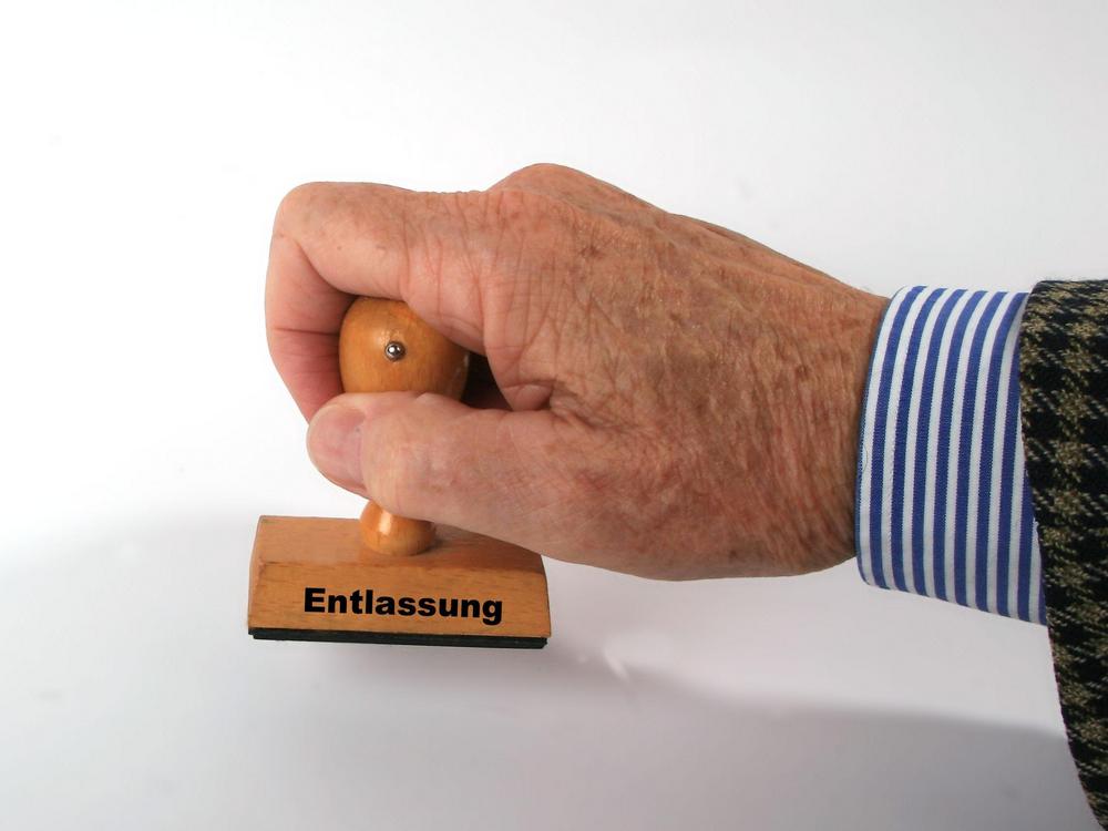 Zu sehen ist eine ältere Hand, die aus der rechten Seite ins Bild kommt und einen mit „Entlassung“ beschrifteten Stempel hält.