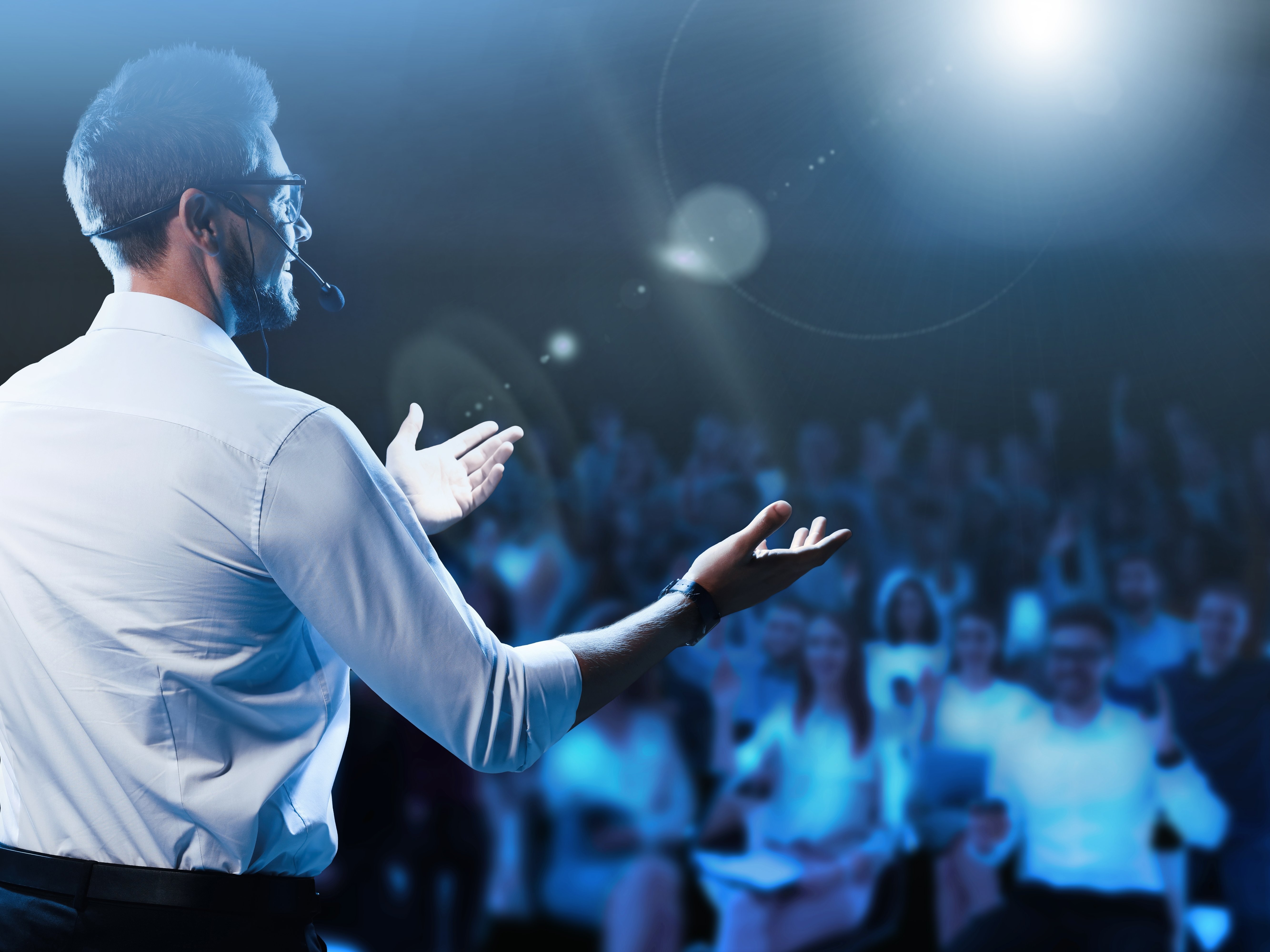 Von hinten ein Mann auf der Bühne, wird mit einem Scheinwerfer bestrahlt. Im Hintergrund dunkel und unscharf ein Publikum