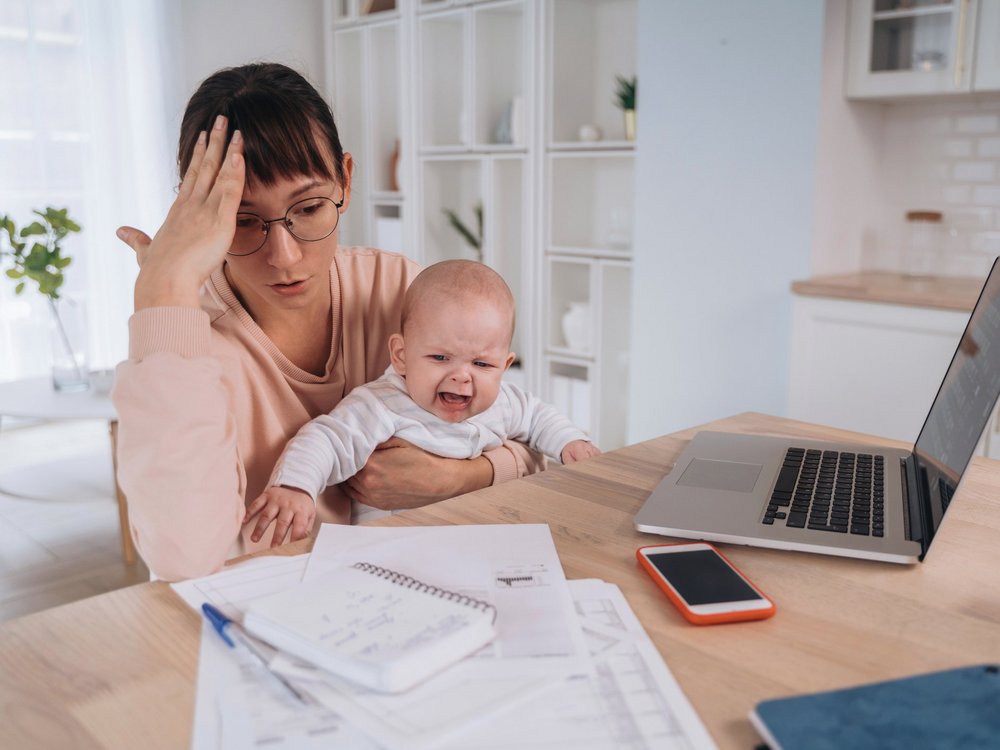 Gestresste Mutter am Schreibtisch vor dem Laptop mit Baby auf dem Arm.