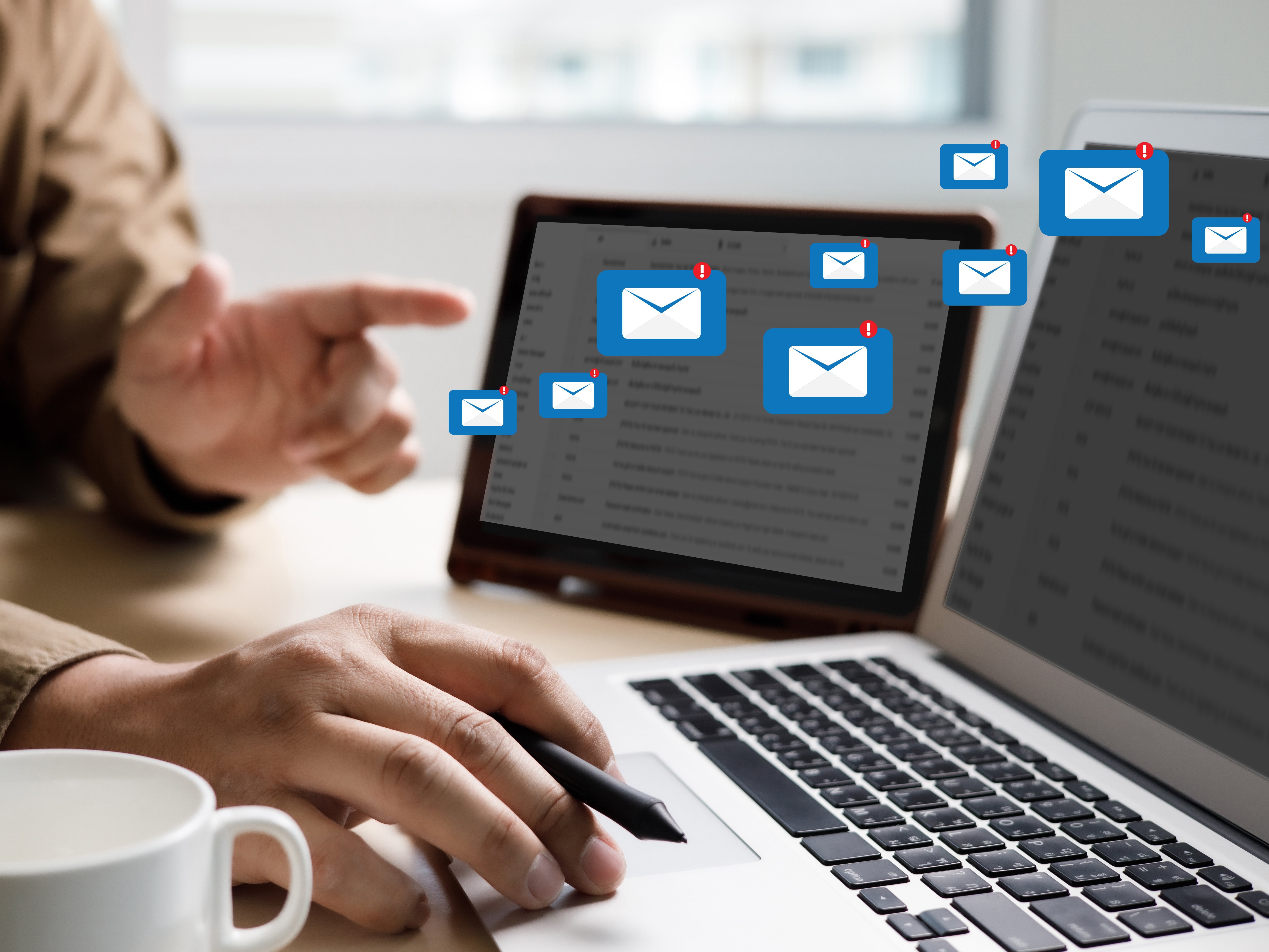 Zwei Laptops nebeneinander. E-Mail Symbole fliegen vom einen Bildschirm zum anderen. Davor jemand, der auf den rechten Bildschirm zeigt.