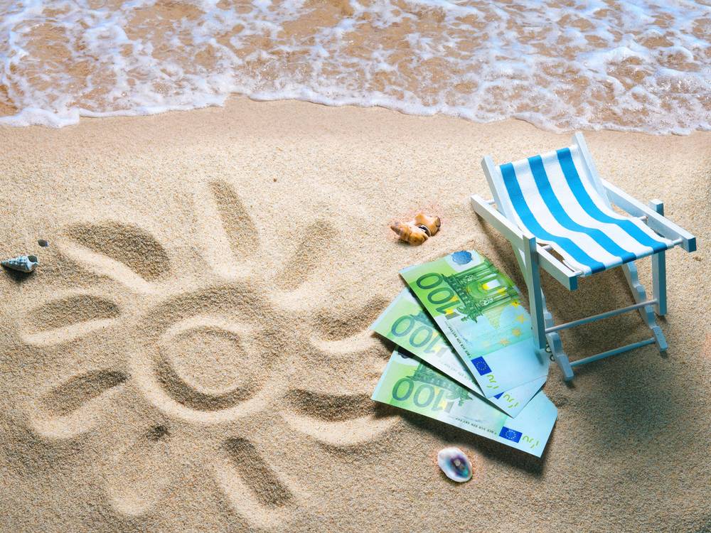 Das Bild zeigt einen Sandstrandabschnitt, auf dem eine Sonne in den Sand gemalt ist. Daneben liegen drei 100-Euro-Scheine. Ebenfalls rechts daneben steht eine Strandliege. In der oberen Bildhälfte ist das Meerwasser zu sehen, das an den Strand gespült wird.