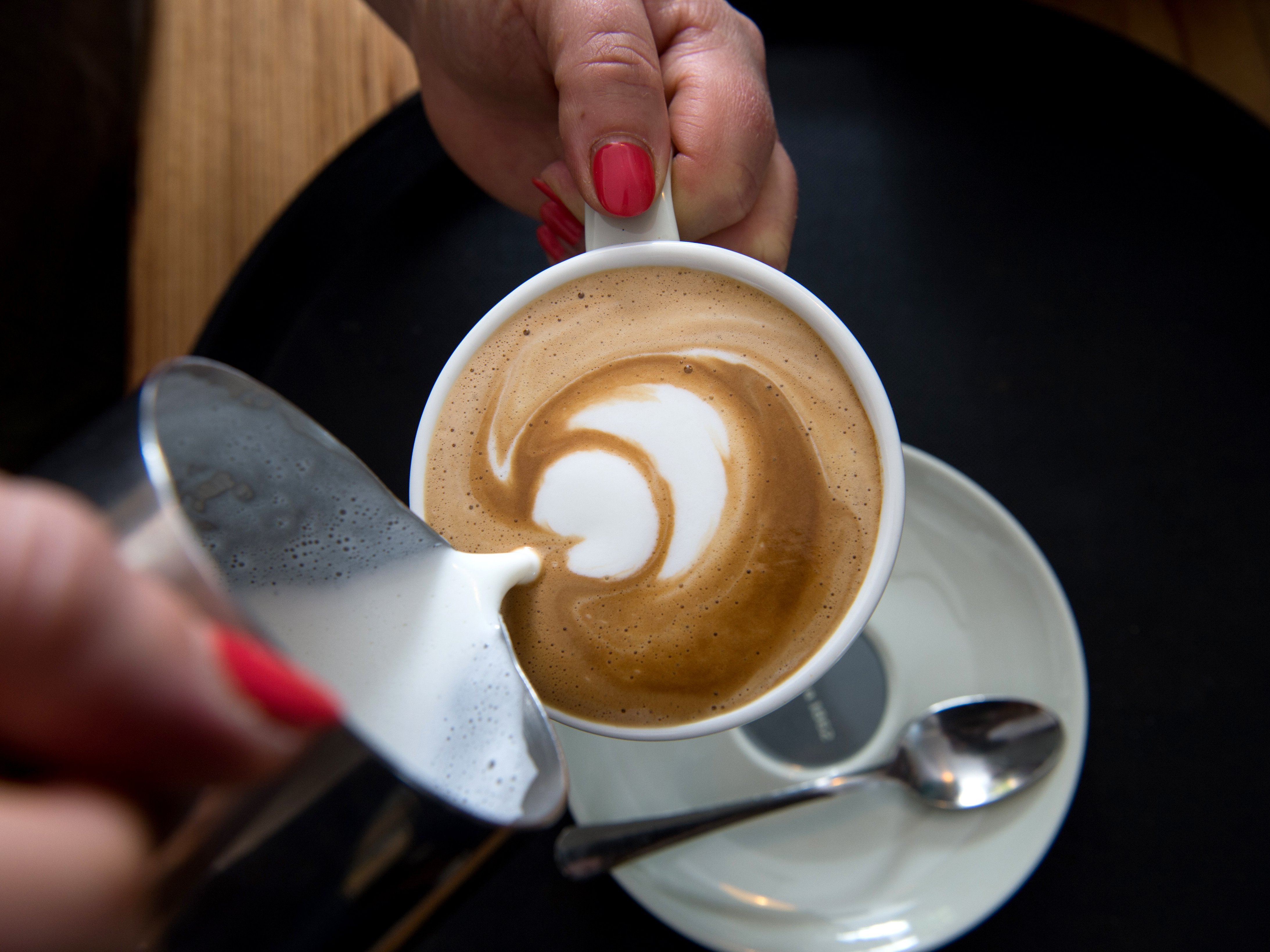 Milch wird aus einer Stahlkanne in eine Kaffeetasse eingeschenkt.
