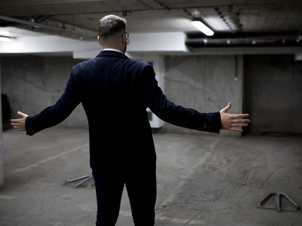 Ein Manager steht in der Tiefgarage vor einem leeren Parkplatz und hat fassungslos die Arme ausgebreitet.