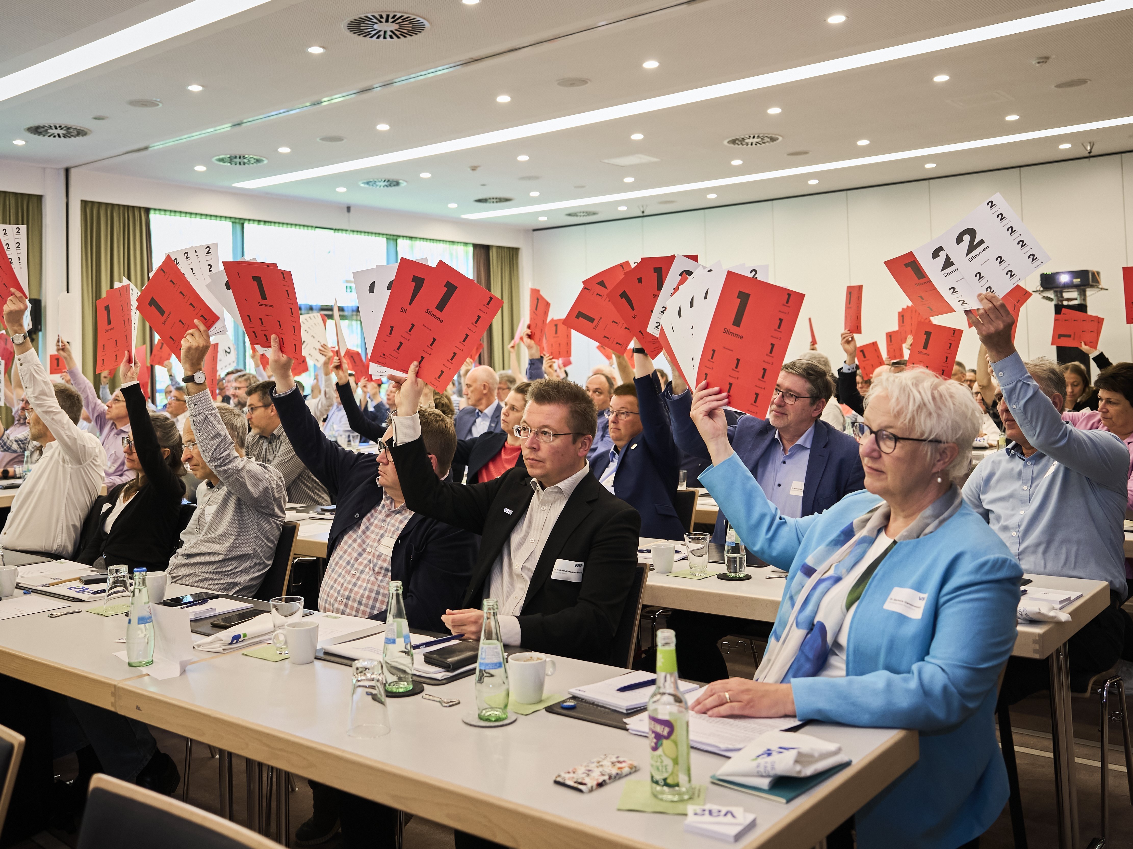 Plenum (Blick nach vorne) hält rote Stimmzettel in die Höhe.