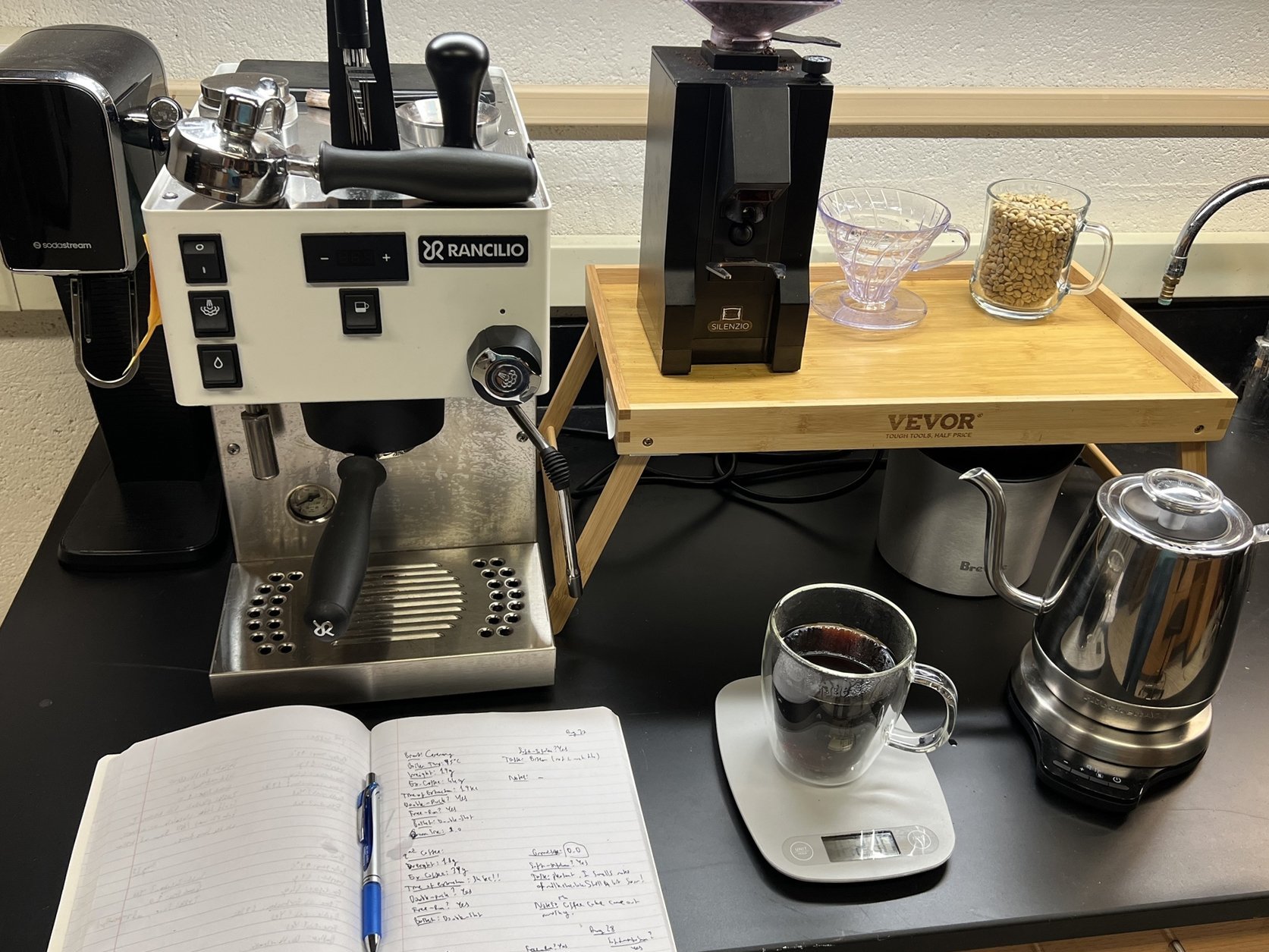 Eine Siebträgermaschine. Davor ein Notizheft mit Notizen. Daneben eine Küchenwaage mit einer Tasse Kaffee darauf. Drum herum viele weitere Utensilien zum Kaffee brühen.