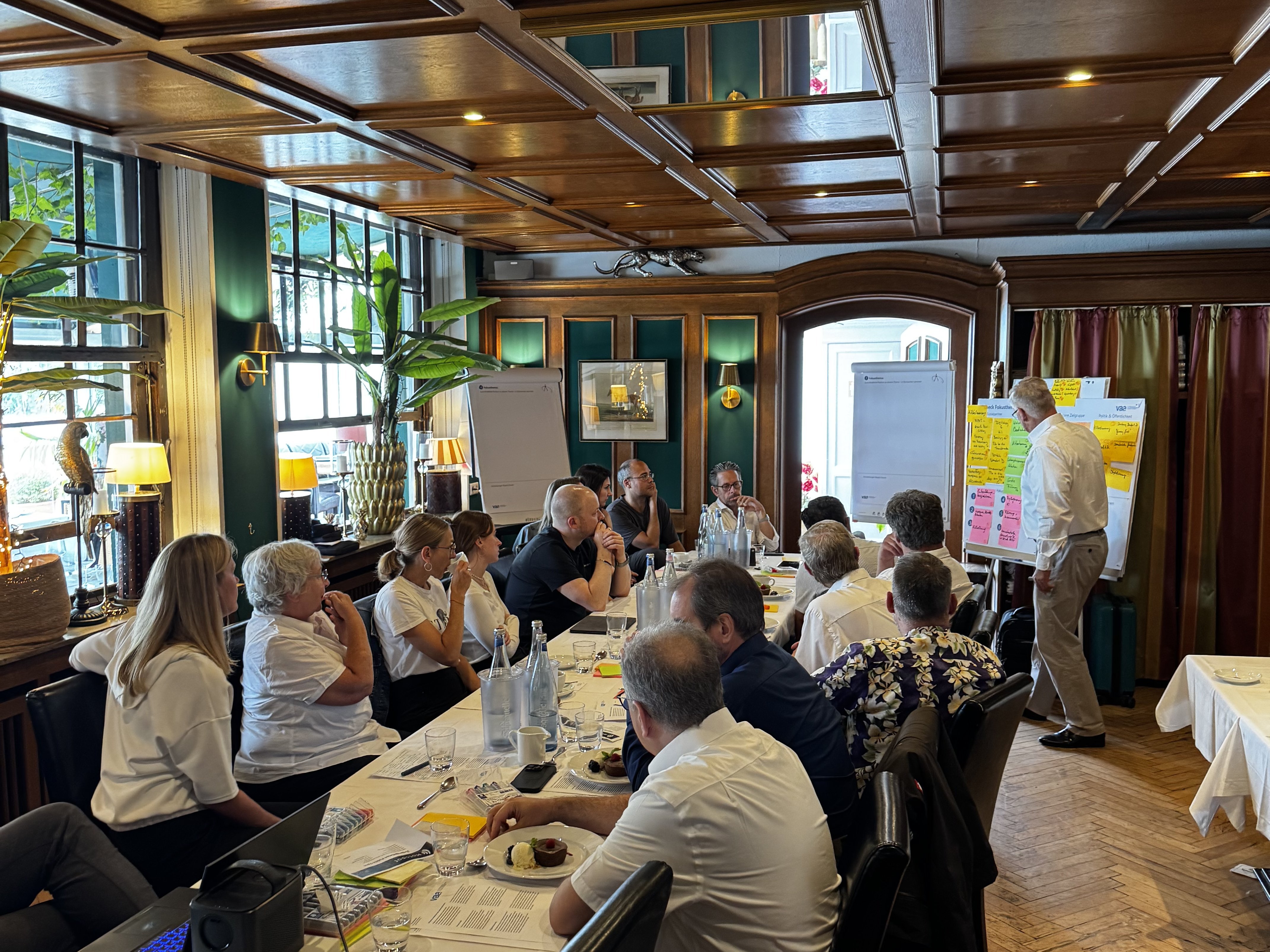 Ein Raum mit großem Tischen, um den die Vorstandsmitglieder und Mitarbeitende der Geschäftsstelle sitzen und in Richtung von Axel Kühn schauen. Er steht an einer Meta-Plan-Wand.