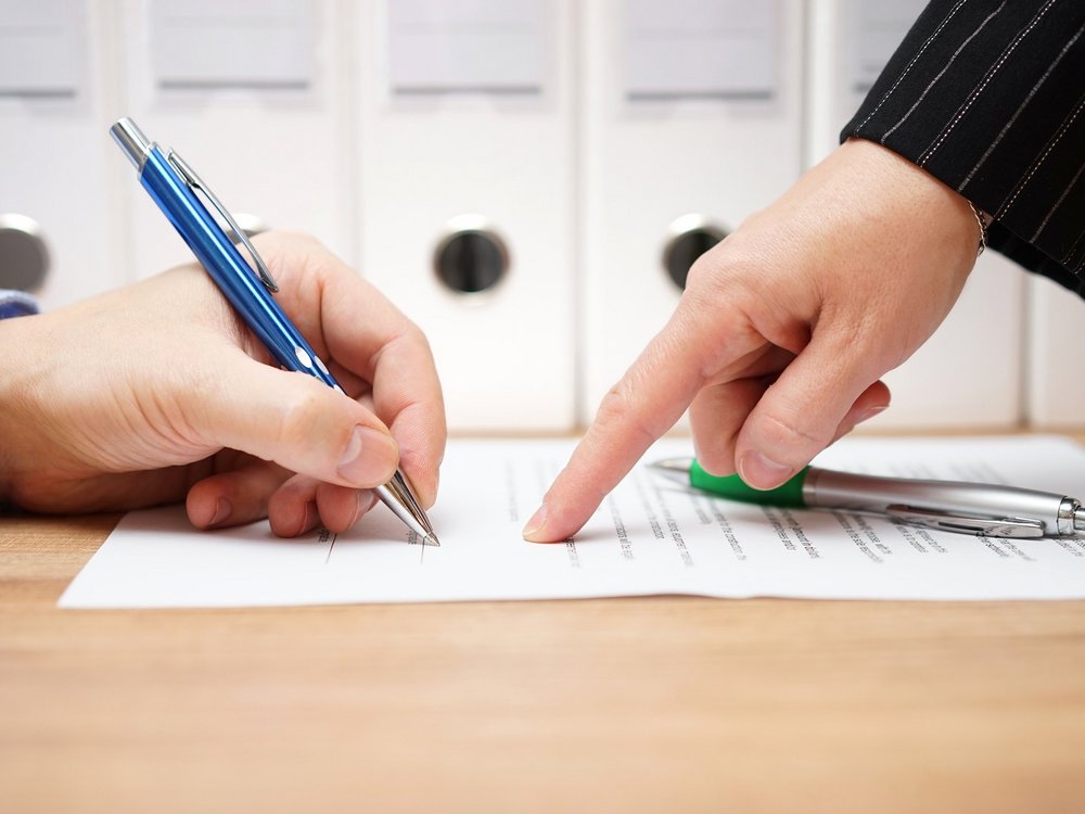Das Foto zeigt zwei Hände über einem Vertrag, der unterzeichnet wird. Die Hand rechts zeigt mit dem Zeigefinger auf die Stelle, an der die andere Hand mit dem Stift die Unterschrift setzen soll.