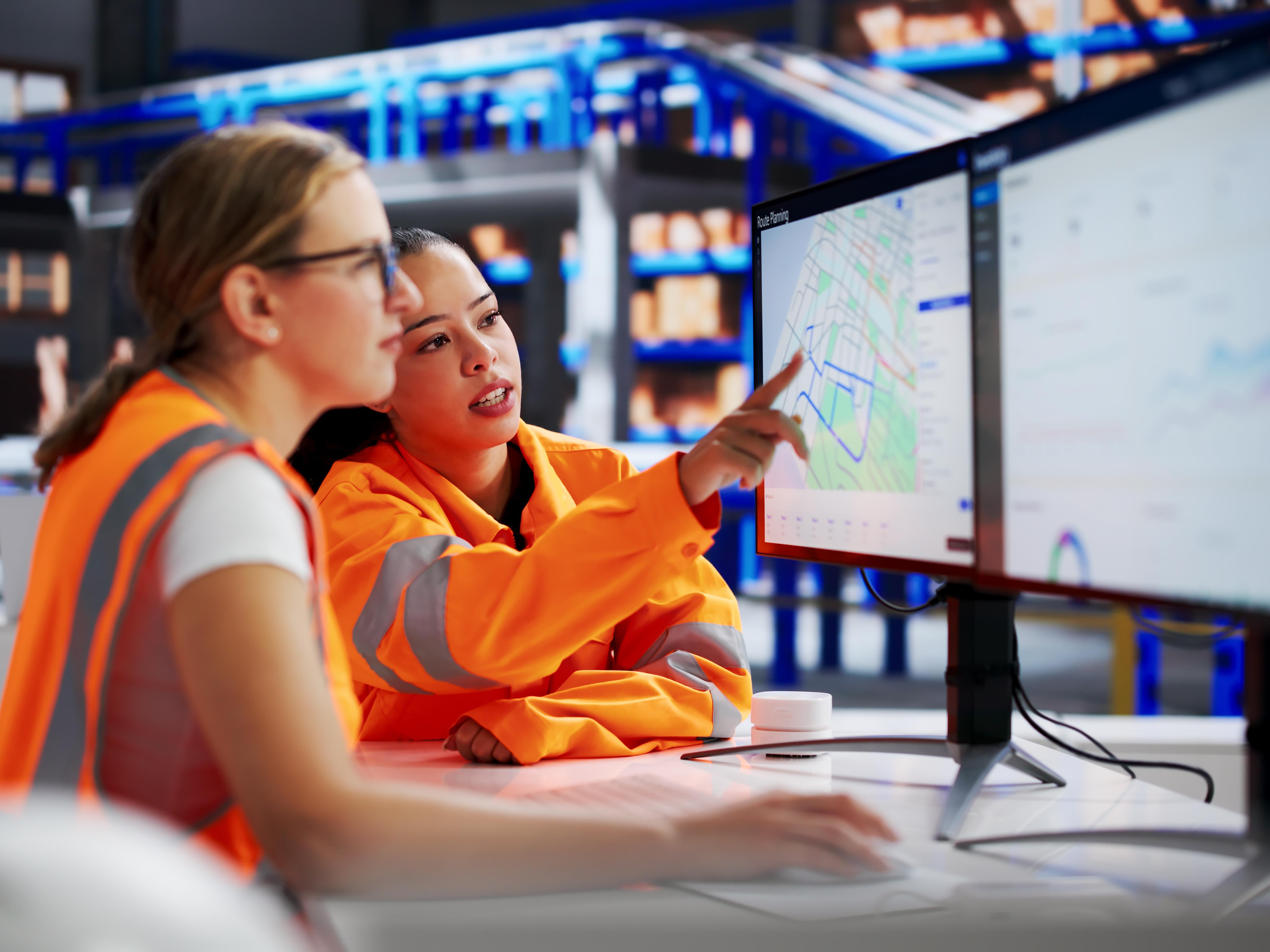 Zwei Personen analysieren Logistikdaten auf Großbildschirmen in einer Lagerhalle.