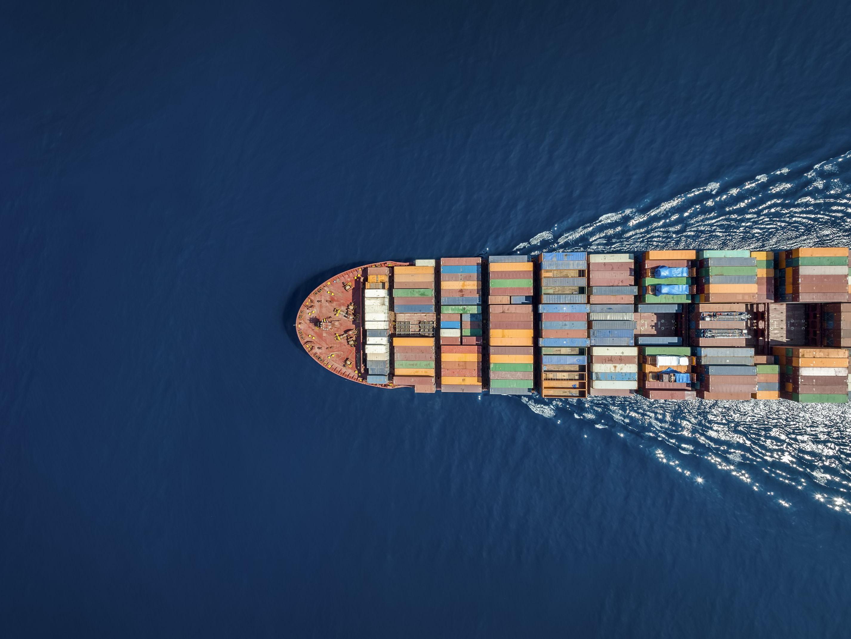 Blick von oben auf ein großes Containerschiff, das von rechts auf dem Meer ins Bild hineinfährt.