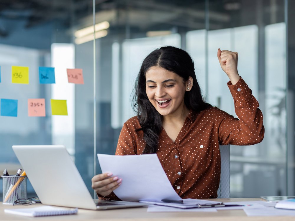 Eine Arbeitnehmerin feiert ihren Erfolg am Arbeitsplatz. Sie hält einen Vertrag in den Händen und hebt freudig die Hand zum Triumphgestus.